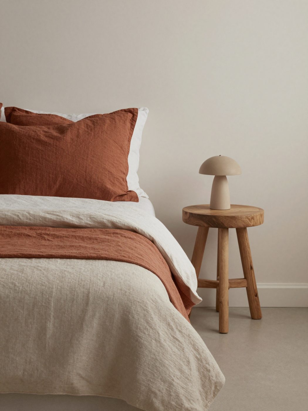 A linen bed with rust cushions, an oak stool and a small lamp.